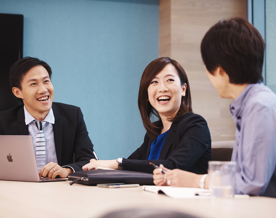Three people sit at a table in a meeting; one uses a laptop, the others hold pen and paper.