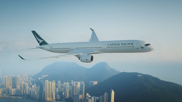 一架國泰航空的飛機飛越高樓林立的城市,背景是山脈和部分多雲的天空。