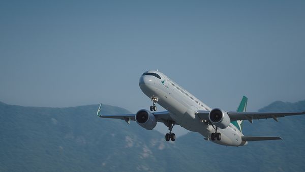一架國泰航空的飛機飛越高樓林立的城市,背景是山脈和部分多雲的天空。
