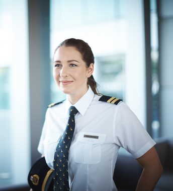 A woman in a pilot uniform stands indoors, looking to the side and smiling.