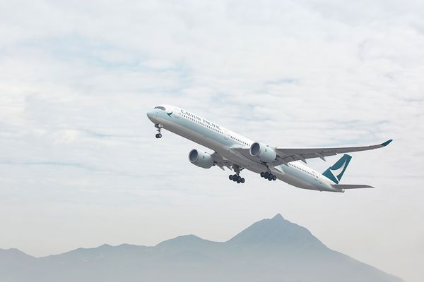A Cathay Pacific Airbus A350 in mid-flight with landing gear partially retracted, set against a cloudy sky and faint mountain outlines.