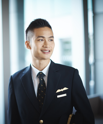 A man in a pilot uniform stands indoors, looking to the side and smiling.