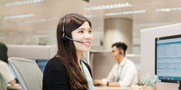 A smiling Customer Care Advisor wearing a headset works at a computer in a modern office. In the background, a colleague focuses on his screen. The atmosphere is professional.