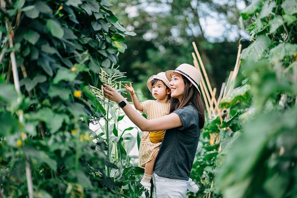 A mother holds her child in a garden, surrounded by greenery. The mother points to the plants while the child looks on, both wearing hats.