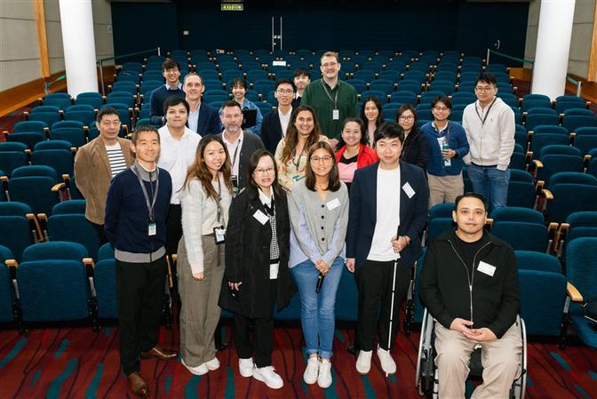 A group of 17 people in an auditorium with blue seats. One person uses a wheelchair, and another holds a white cane.