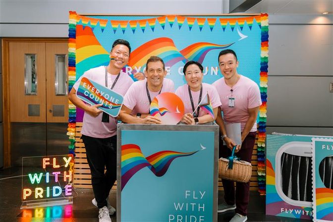 Four people stand behind a flight trolley with a vibrant rainbow-themed backdrop and neon sign featuring the slogan “FLY WITH PRIDE”.