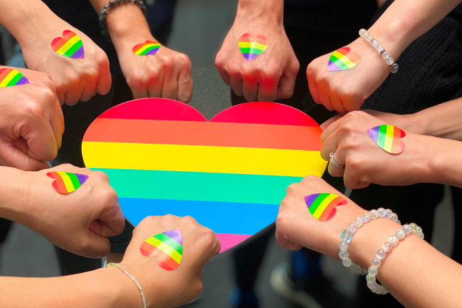 A group of hands with rainbow heart stickers on their fists surround a large rainbow-colored heart on a table, symbolizing support for LGBTQ+ pride.